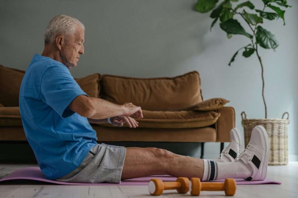 Elderly man in blue shirt exercising on a yoga mat indoors with dumbbells in view.